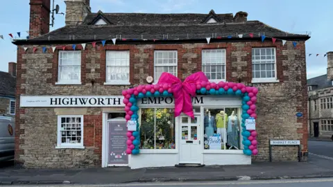 Aga Marsden A shop decorated with a large pink bow with blue and pink balloons outlining the shop front. The words "Highworth Emporium" can be seen beneath the bow.