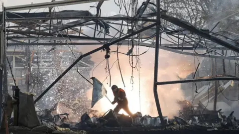 A rescue worker in the middle of rubble at a commercial building where only mangled metal frames remain; fire and smoke are shown in the background in the huge void betwen buldings