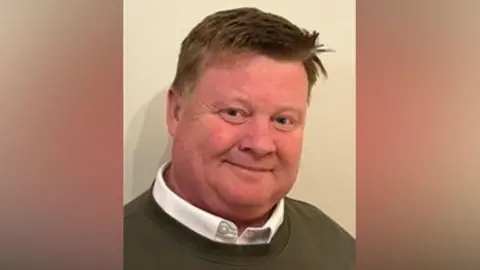 Warwickshire County Council Conservative councillor and council chairman Dale Keeling looks at the camera. He wears a green jumper and white unbuttoned shirt