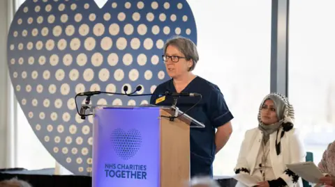 NHS Charities Together A woman with short grey hair wearing dark blue medical scrubs stands at a podium which says 'NHS Charities Together'. A woman wearing a grey hijab and white clothes sits behind her.