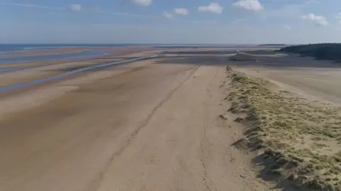 Martin Barber/BBC Drone shot of a wide-open beach with an expanse of sand, low sand dunes and pine woods. The sea is blue, as is the sky, and there are fluffy white clouds.