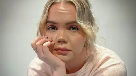 Meri-Tuuli Auer looking towards the camera, she is seated with her head resting in her hand. She has blond hair and is wearing a light pink short sleeved woollen top.