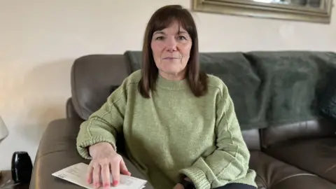 BBC A woman with shoulder-length dark hair, wearing a green jumper, sits on a sofa looking at the camera