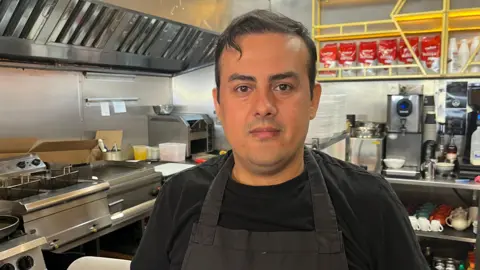 Jonny Humphries/BBC Ozy Kahraman, who has short black hair and is clean shaven, stands in the kitchen area of a cafe, wearing a black t-shirt under a black apron. 