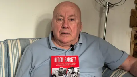 An old man stares at the camera wearing a blue polo and holding a memoir