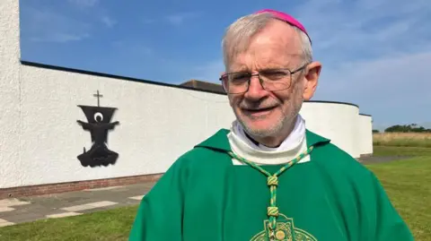 A bishop wearing a green cassock with a white neckline and he is wearing a purple skull cap. He has clear glasses and is smiling into the camera. The wall surrounding a church is in the background, and there is a pathway around it. Green grass is also visible in the background.