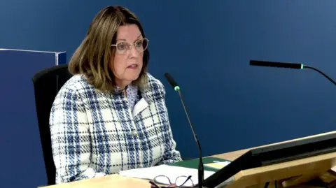 Agnes McCusker sits at a desk and speaks into a microphone during the inquiry. She wears a cream and navy tartan jacket, has shoulder-length brown hair and wire-framed glasses.  