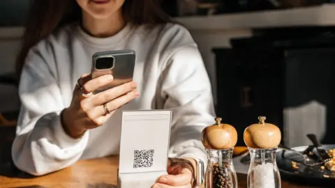 A woman sat at a restaurant table scanning a QR code with her phone.There is a salt and pepper shaker and a half eaten plate of food next to her on the table