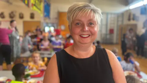 Hayley Clarke / BBC Head teacher Clare Morton smiling at the camera, with children eating their school dinners in the lunch hall behind her. Clare has short, blonde hair with a fringe, and is wearing a black dress and red lipstick.