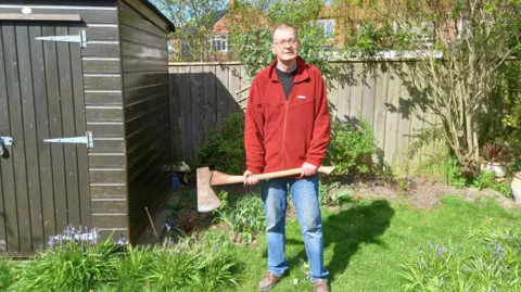 Ian Fraser A man in his 60s wearing jeans and a dark red fleece is standing in a garden next to a shed and holding a garden axe 