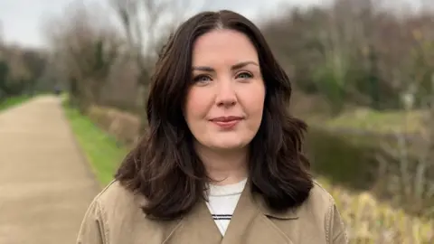 Rebecca Harper wearing a brown coat and a white jumper looking at the camera. She is standing outside and there is a path, some grass, trees and a small body of water in the background