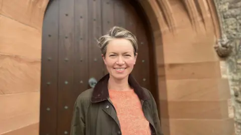 A woman with short brown hair, wearing an orange top and brown jacket, stands in front of a church door