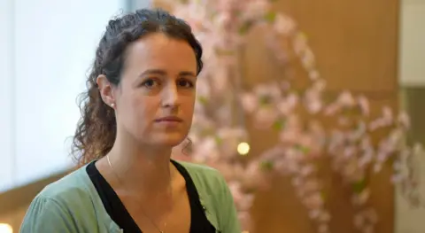 Rebecca Pursey is looking seriously at the camera. She has dark brown curly hair tied back in a ponytail, and is wearing a green cardigan with a black top underneath.