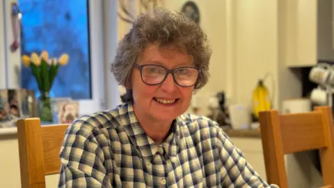 A woman with curly grey hair and black glasses is sitting in a kitchen and smiling at the camera. She is wearing a blue and white checked shirt.