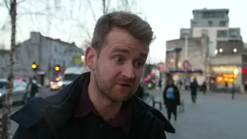 A man in a dark jacket. He has a sandy-coloured cropped beard and dark-blond hair