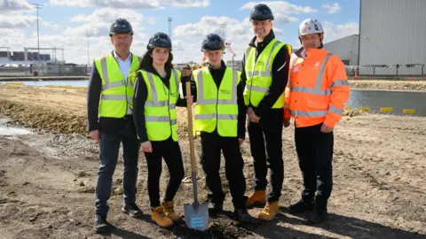 Uber Boat by Thames Clippers Several people in high-visibility jackets stand on a construction site. The two in the middle of the frame both have a hand on a spade which is pointed into the ground. 