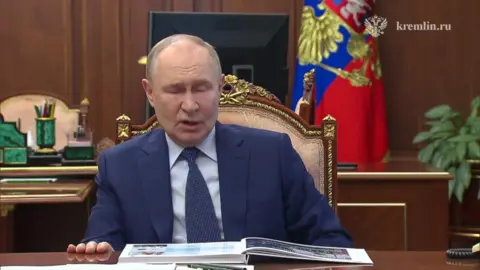 Putin, in a blue suit, shirt and tie, speaks from an ornate chair at a desk in the Kremlin