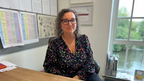 Woman wearing a multi- coloured blouse, glasses, looking at the camera in an office setting