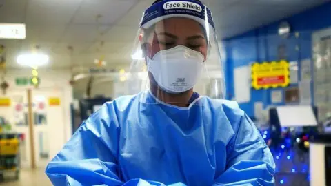 An NHS working in blue overalls wearing full PPE in a hospital setting. 