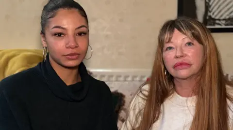BBC Jayne and Christiana pictured next to each other looking at the camera. Christiana on the left has her dark hair tied back and is wearing silver hoop earrings. She is wearing a black knitted jumper. Jayne has long light brown hair and a fringe. She is wearing a white knitted jumper.