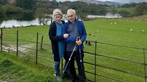 A woman and a man are standing alongside a metal fence with a field and large body of water on the other side. There are sheep in the field and in the distance is woodlands and further in the distance is a range of hills.