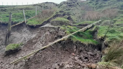 Cyngor Gwynedd A grassy hillside with mud where a sinkhole has appeared on a pathway