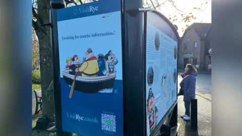 Signs encouraging people to visit Rye. A group of cartoon men can be seen paddling in a boat. 