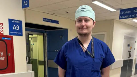 Royal Wolverhampton NHS Trust A man in blue theatre scrubs standing in a hospital corridor. He is smiling.