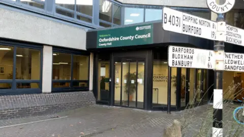 BBC Oxfordshire County Council's County Hall in Oxford. The picture shows the front doors of the building, with a green sign above that says "Welcome to Oxfordshire County Council County Hall". There is a black and white sign to the right of the picture pointing out distances to other places in Oxfordshire.