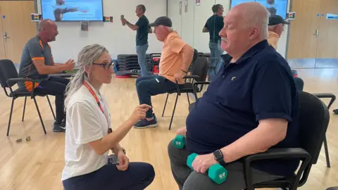 Simon Thake A woman with long blonde hair crouches down to encourage an older man sitting in chair with small green weights. In the foreground other men of the gym other men do stretching exercises