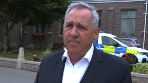 BBC A man with short grey hair and wearing a black sit and white shirt is pictured outside a granite parish hall. A police car is visible behind him, along with some plants.