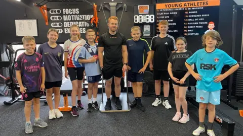 BBC Eight children stand in a gym. They are all dressed in sports gear, including trainers, shorts and sports shirts. A man stands in the middle of them with red hair, a black t-shirt, shorts and trainers. Gym machines can be seen in the background with a motivational slogan on the wall, a timer and a list of fitness activities displayed on a large screen.