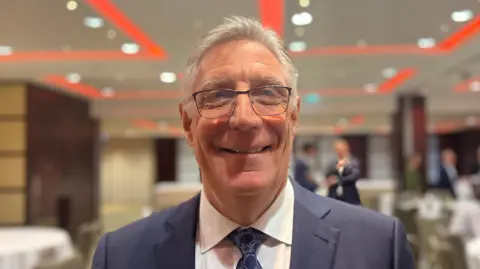 BBC Malcolm is a man with white hair. He is wearing a navy suit with a white shirt and navy patterned tie. He is wearing glasses. He is stood in a conference room and is smiling at the camera.