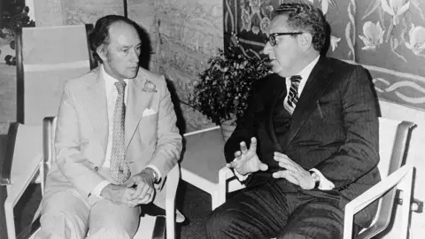Getty Images A black and white image showing Pierre Trudeau and Henry Kissinger in conversation, seated in chairs side by side. 
