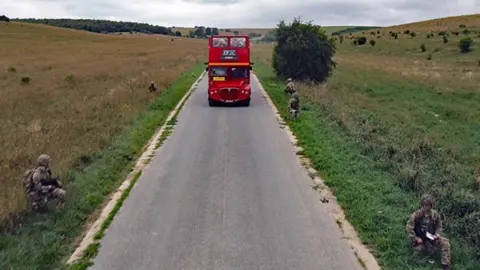 ianVisits A red double-decker Routemaster bus travels along an unmarked road. Passengers can be seen sitting on the upper deck of the bus, which has an open top. The road is flanked by five soldiers wearing camouflage uniforms and kneeling in the grass verges