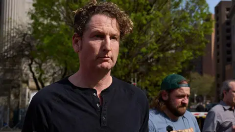 Getty Images White man with curly blonde hair and black henley shirt stares off unsmiling in front of a green tree and man with backward-turned green baseball cap