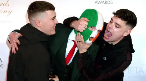 PA Media Members of Kneecap, Liam Og O Hannaidh (Mo Chara), JJ O Dochartaigh (DJ Provai), and Naoise O Caireallain (Moglai Bap) attending the Irish Film and Television Academy Awards Ceremony. They have their arms round each other.
