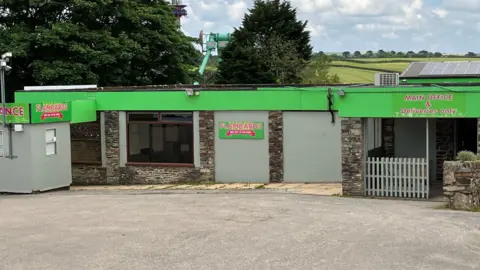 The main office of Flambards with green hoardings along the top of a single-storey building with grey walls, a couple of windows and signs with Flambards written on them in pink letters.