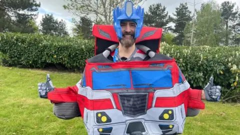 James Burridge/BBC Matt Batchelor, with two arms held out, dressed in a costume, as Optimus Prime. He has a beard and is standing on grass with buses behind him. 