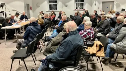 Around 50 members of the public are sat on five rows of black plastic chairs facing a row of four people sat at a table. Many of those in the hall for the public meeting are elderly.