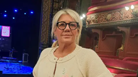 Adele Teale A woman with blonde hair and glasses stands in an empty theatre.