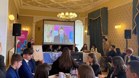 Around 20 people are sitting at tables in a conference room. One girl is standing at a podium. She is being shown on a projector screen in the middle of the room.