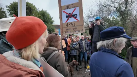 A crowd holding placards listen to a speaker standing on a footstool by the banks of the chalk stream