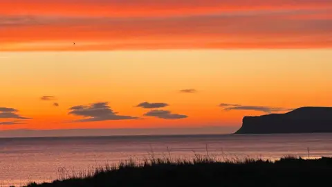 Superted Marske-by-the-Sea under a sunrise. The skies are shades of orange, yellow and burnt amber, with a few scattered grey clouds in a line of them on the horizon. The sea is a darker shade of orange-grey. There is a silhouette of cliffs to the right, with a grassy bank in the foreground.