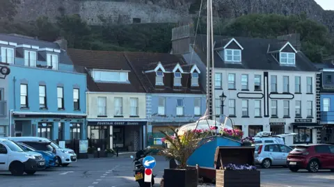 A picture of shops in Gorey Harbour. The bottom of Mont Orgeil Castle is visible on the cliff face above.