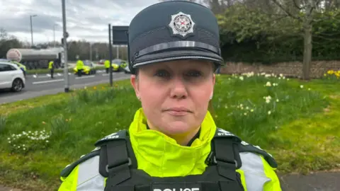 A police officer is wearing a high viz jacket. Her body protector says police. Her hat has the PSNI emblem.