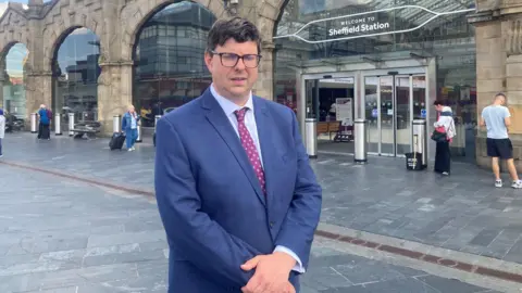 BBC/Naj Modak A man with dark hair and glasses is wearing a blue suit, pale blue shirt and maroon tie. He is standing outside Sheffield railway station which has large glass windows and stone arches