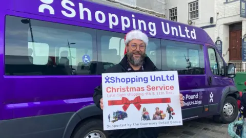 A man with a grey beard, glasses and a Santa hat, stood in front of a purple van with the words 'Shopping UnLtd" printed onto it, holding a sign with the same text.