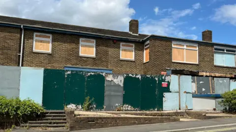 A street view of a derelict building with all of the windows boarded up. A wooden barrier has been put up around the base of the building and it is very worn.