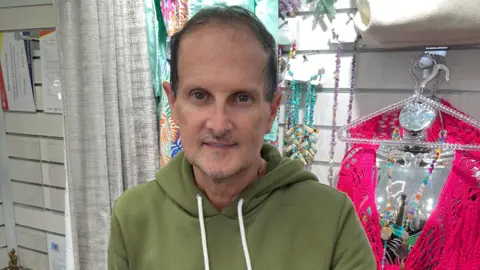 LDRS A man with short dark hair, wearing a green hooded top, stands in a clothes store with items of clothing hanging on the wall behind him.